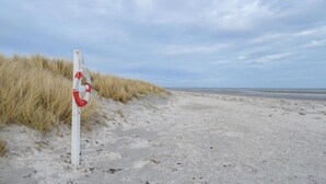 Una playa arenosa con un flotador rojo sujeto a un poste blanco, rodeada de hierba alta.