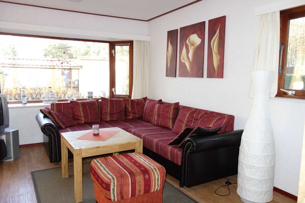 A living room with a sectional sofa, a small table, a black leather armchair, and a tall white lamp. There are three paintings on the wall and a window with curtains.