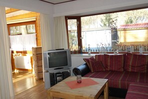 A cozy living room with a TV, wooden furniture, and a large window with a view of the sea and trees.