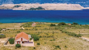 En ejendom ved kysten med et hus og rødt tag, omgivet af græslandskab og udsigt til havet samt fjerne bjerge.