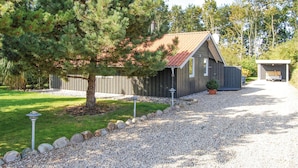 A rustic cabin with a gravel driveway, surrounded by trees and a fenced yard.