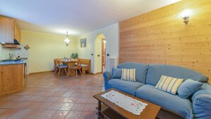 Un soggiorno con un divano blu, pareti rivestite in legno, un'area pranzo con tavolo e sedie, e una cucina con mobili in legno e pavimento piastrellato.