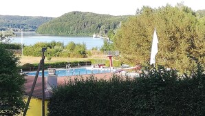 A swimming pool with diving boards, surrounded by trees and a view of a lake and hills.