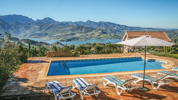 Ein rechteckiger Swimmingpool mit klarem, blauem Wasser, umgeben von einer gefliesten Liegefläche. Liegestühle und Sonnenschirm sind vorhanden. Im Hintergrund sind Berge und ein Tal mit einem See zu sehen.