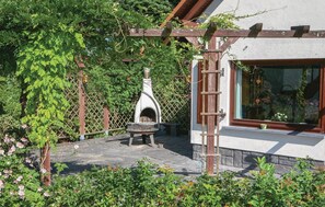 Une terrasse en pierre avec un four à bois, entourée d’une végétation luxuriante et agrémentée d’une pergola en bois.