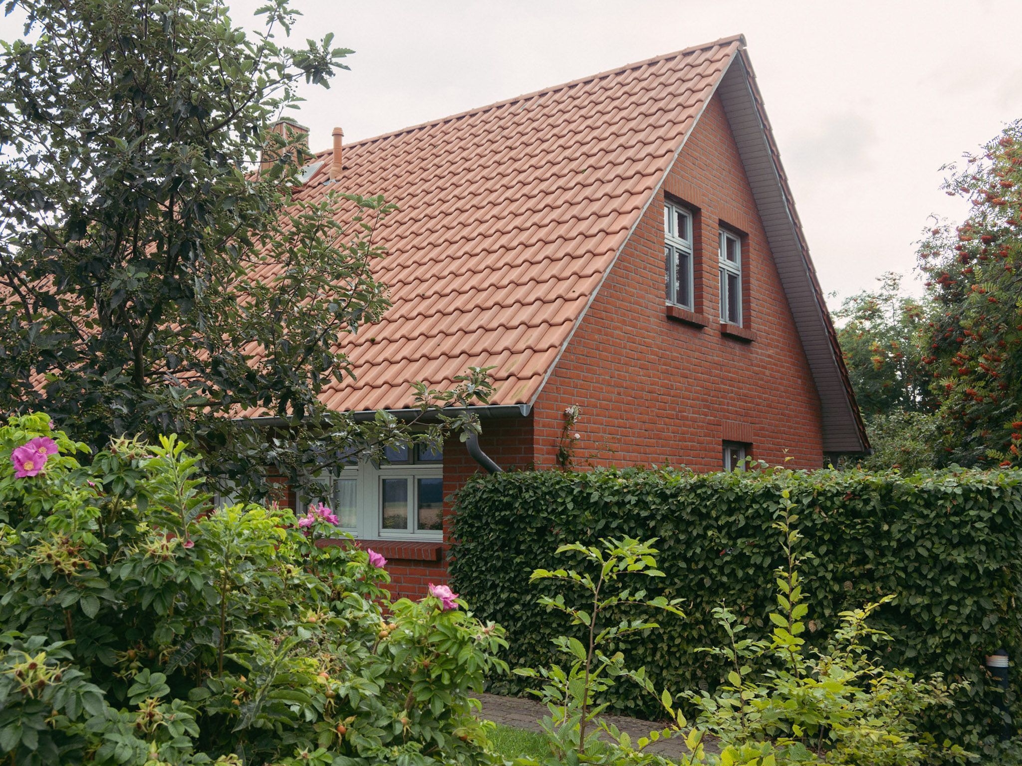 A red brick house with a sloped roof, surrounded by lush greenery and flowering plants.