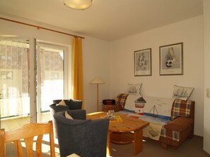 Ein Wohnzimmer mit einer Couch, einem Holztisch und einem Sessel. Ein Fenster mit Vorhängen, eine Lampe und zwei gerahmte Bilder sind an der Wand zu sehen.