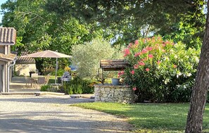 Une terrasse avec une table et des chaises, une structure en pierre avec un banc, et un jardin agrémenté de fleurs roses.