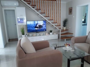 A living room with a TV, two sofas, a glass coffee table, a staircase, and a kitchen in the background.