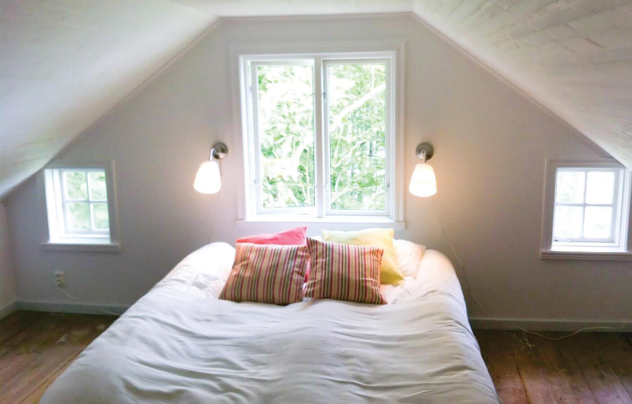 A bedroom with a sloped ceiling, a large bed with white bedding, two wall-mounted lamps, and two windows with a view of greenery.