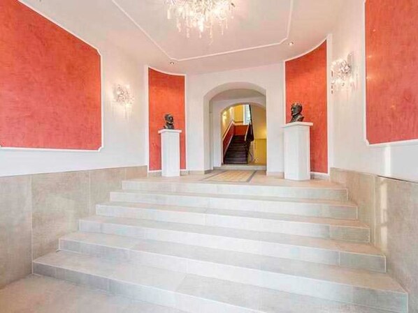 A grand staircase with marble steps leading to an upper level, flanked by red walls and white columns with busts.