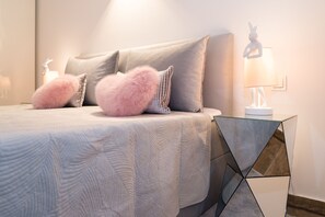 A neatly made bed with a textured white duvet, two pink fluffy pillows, and a geometric glass side table with a white lamp.
