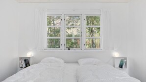 Une chambre avec une grande fenêtre, deux tables de chevet avec des livres, et un lit avec une literie blanche.