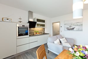 A modern kitchen with white cabinetry, a built-in oven, and a microwave. There is a dining area with a wooden table and a grey sofa.