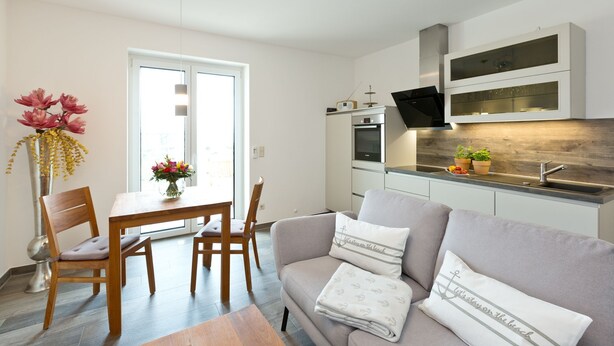 Una cocina moderna con horno y microondas integrados, un comedor con mesa y sillas de madera, y una sala de estar con sofá y cojines decorativos.