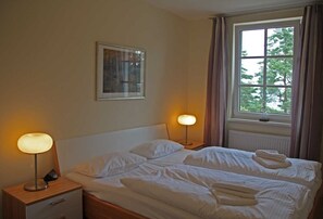 A neatly made bed with pillows, a bedside lamp, a nightstand, a framed picture on the wall, and a window with curtains.