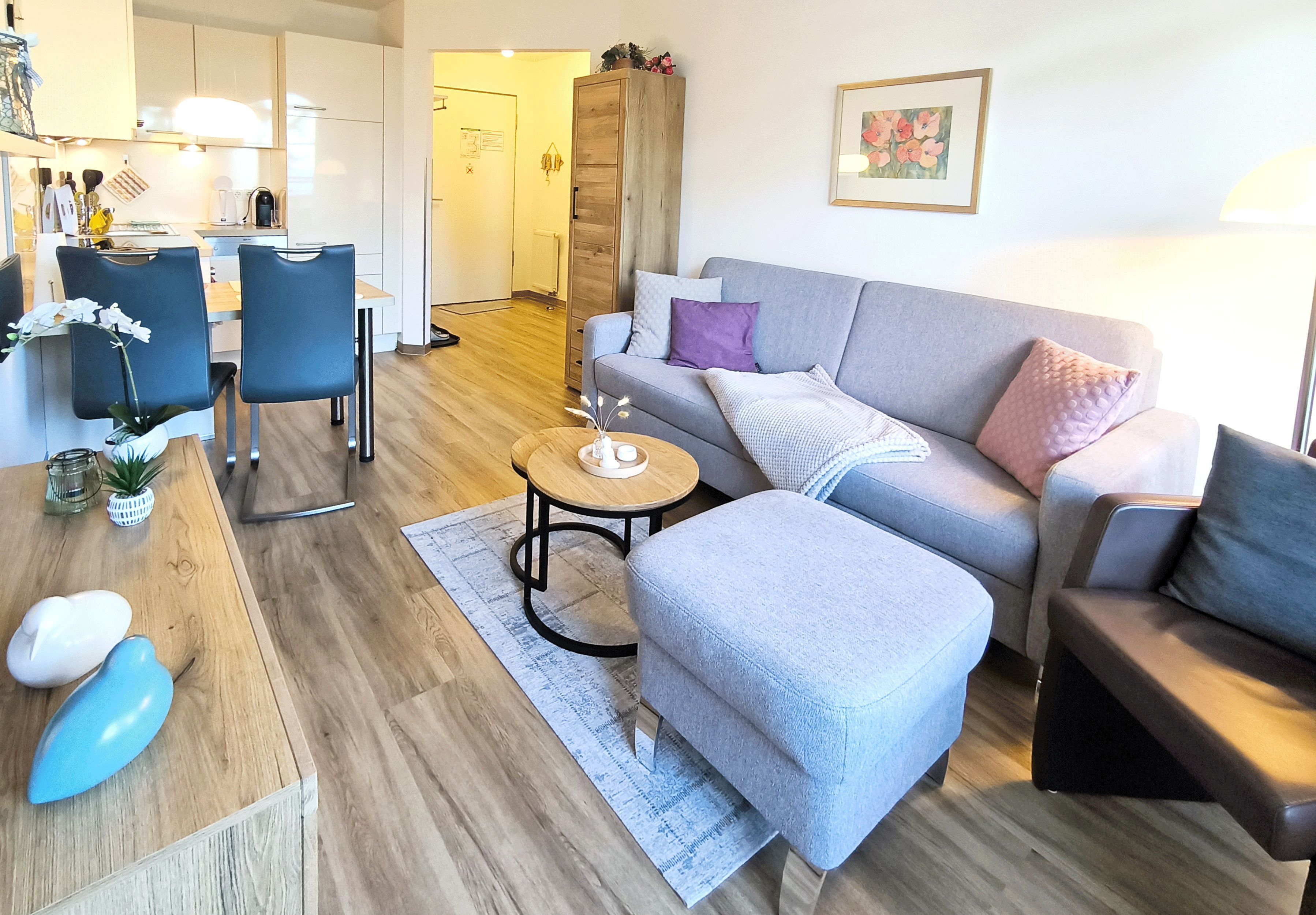 A modern living room with a grey sofa, ottoman, and chairs, a wooden coffee table, and a kitchen in the background.