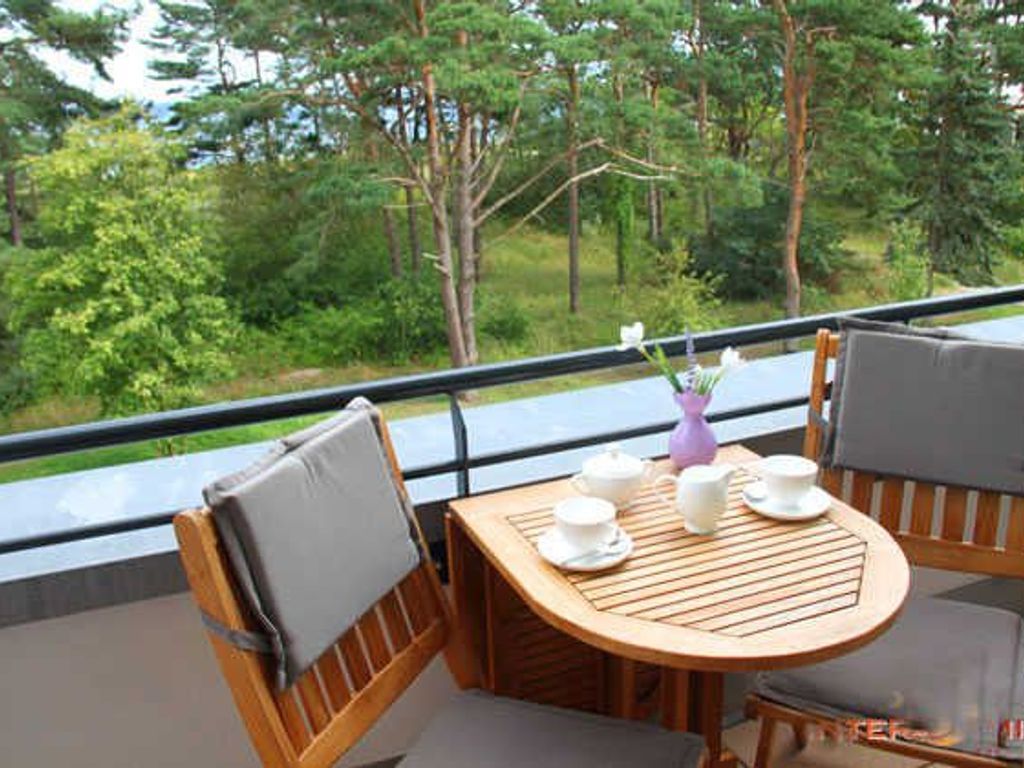 Een balkon met een houten tafel en stoelen, een bloemenvaas en uitzicht op een bos.