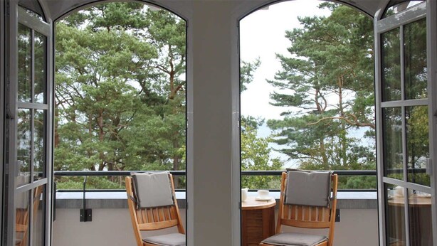 A balcony with two wooden chairs and a small table, overlooking a forested area.