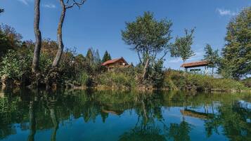 A serene lakeside setting with a wooden cabin and a gazebo, surrounded by lush greenery and trees reflecting in the calm water.