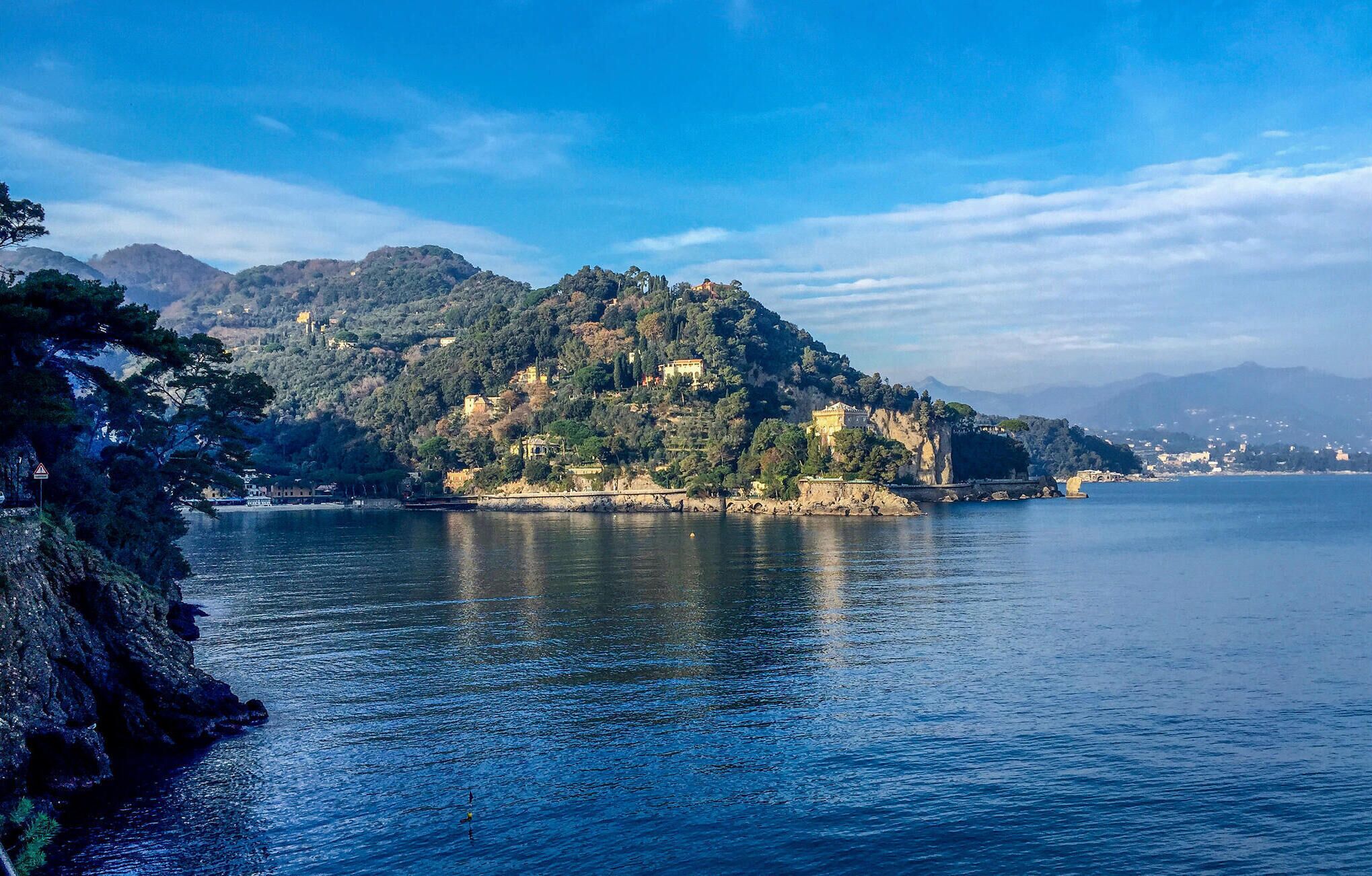 Paesaggio costiero con una collina, edifici, un mare calmo e montagne in lontananza.