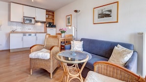 Un espace de vie compact comprenant un coin cuisine, un coin repas et un coin salon avec un canapé bleu et des fauteuils en osier.