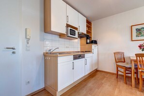 A compact kitchen with white cabinets, a microwave, and a coffee maker. There is a wooden dining table with chairs and a framed picture on the wall.