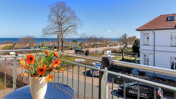 Un balcon orné d’un pot de fleurs, une vue sur une route où circulent des voitures et un bâtiment coiffé d’un toit recouvert de tuiles rouges.