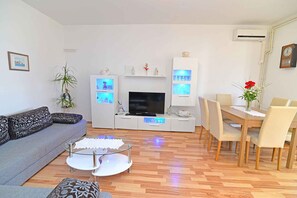 Een moderne woonkamer met een grijze bank, een glazen salontafel, een flatscreen televisie, een eettafel met stoelen en decoratieve wandplanken.