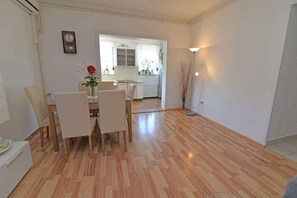 Een eetgedeelte met houten vloer, witte stoelen, een tafel en een vaas met rode bloemen. Op de achtergrond is een keuken zichtbaar.