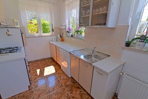Een functionele keuken met een gasfornuis, een spoelbak en een vaatwasser. Op de vensterbank staan potplanten en er is een kastje boven de spoelbak.