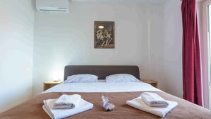 Hotelkamer met een bed, twee kussens, een nachtkastje, een lamp, een ingelijste foto aan de muur en een raam met gordijnen.