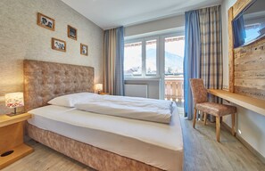 Une chambre d’hôtel avec un grand lit, un bureau en bois, une chaise et une vue sur les montagnes par la fenêtre.