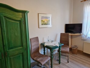 Una habitación con un armario verde, una pequeña mesa con sillas, un cuadro en la pared, un televisor sobre un mueble bajo y una ventana con cortinas de encaje.
