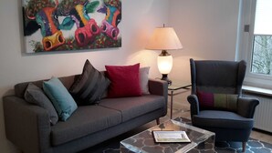 Un soggiorno con un divano grigio, una poltrona nera, un tavolino da caffè in vetro e un quadro astratto colorato appeso alla parete.