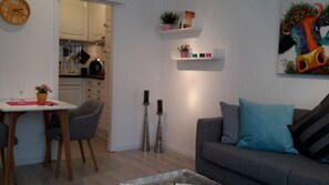 Ein modernes Wohnzimmer mit einem grauen Sofa, einem Esstisch mit Stühlen und einem durchgehenden Gang sichtbaren Küchenbereich.