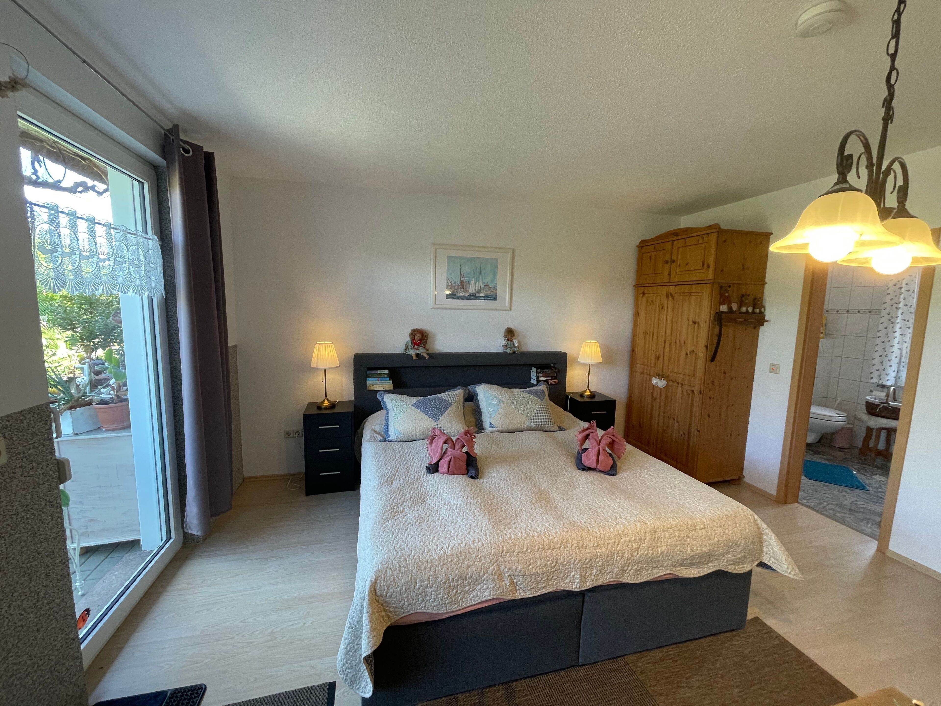 Une chambre à coucher avec un lit, deux lampes de chevet, une table de nuit, une armoire en bois et une salle de bain visible à travers une porte ouverte.
