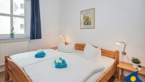 A hotel room with a wooden bed, white linens, a window with curtains, a painting on the wall, and a small plant on the nightstand.