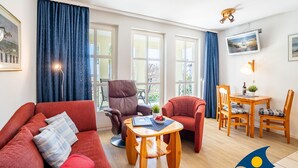 A living room with a red sofa, a brown armchair, a wooden coffee table, and a dining table with chairs. There are large windows with blue curtains, a ceiling fan, and a mounted TV.