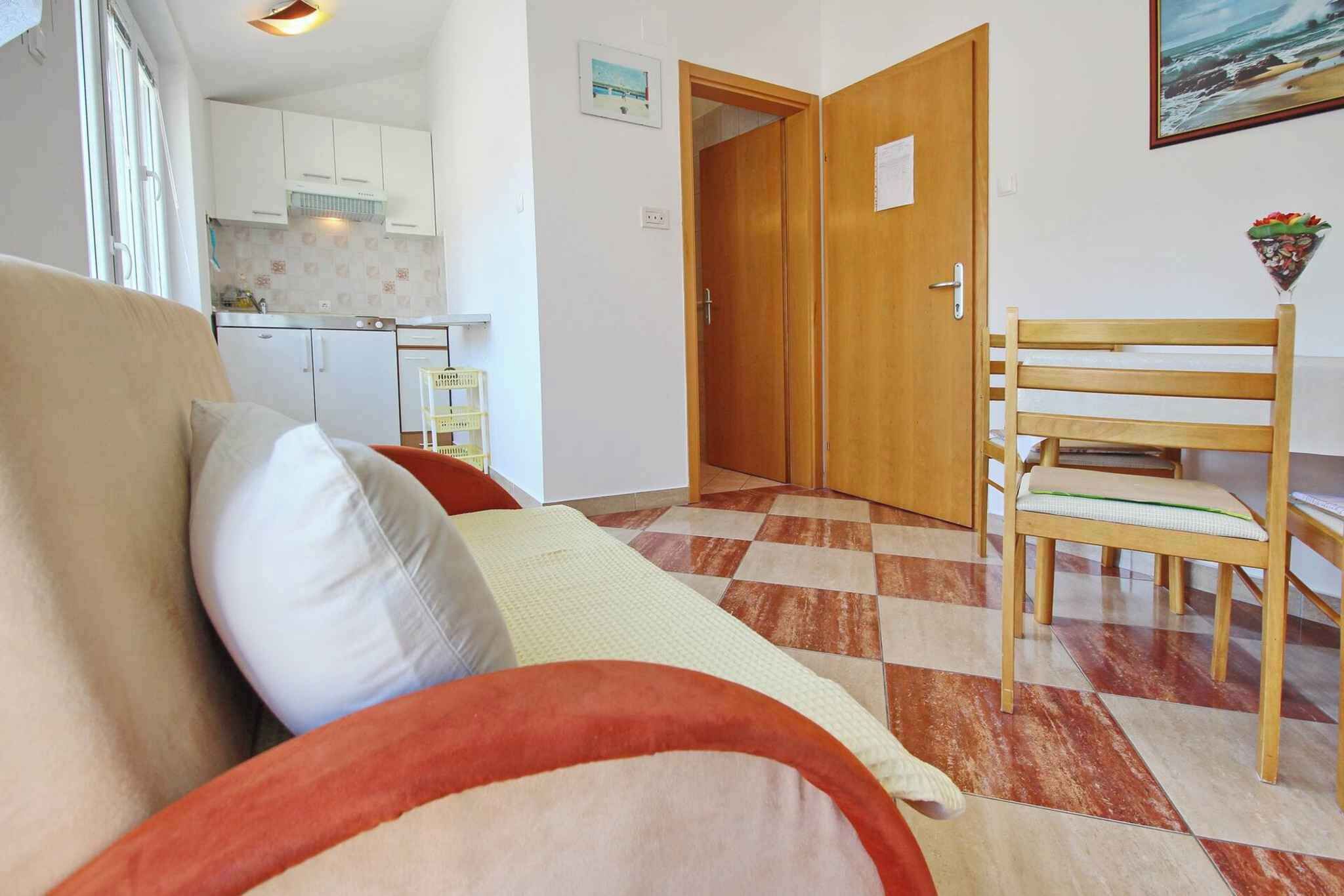 Une pièce avec un canapé, une chaise en bois, un sol à carreaux, une cuisine à l’arrière-plan et une porte avec une pancarte.