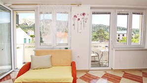 Une pièce comprenant un lit, une fenêtre donnant sur des bâtiments et des collines, et une porte menant à un espace extérieur.
