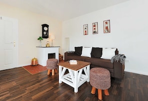A living room with a brown sofa, white cushions, a wooden coffee table, and two wicker ottomans. There is a fireplace with a clock above it and three framed pictures on the wall.