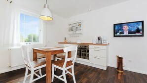 Eine moderne Küche mit weißen Schränken, einem Holzespeisetisch und Stühlen. An der Wand ist ein Fernseher angebracht, der eine Nachrichtensendung zeigt.