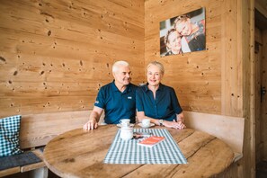Um interior de madeira com uma mesa redonda de madeira, duas cadeiras, toalha de mesa xadrez, uma fotografia emoldurada na parede e um banco com almofada xadrez.