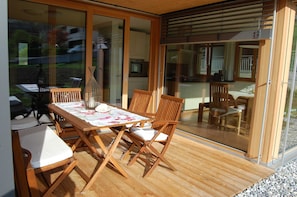 Una terraza de madera con una mesa y sillas, una puerta de vidrio que da a una cocina y vistas al exterior.
