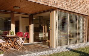 Ein modernes Haus mit einer Holzterrasse, Schiebetüren aus Glas und einem überdachten Außenbereich zum Sitzen.