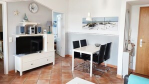 Uma sala de estar com televisão, mobiliário branco, uma área de jantar com mesa e cadeiras, um relógio na parede e uma imagem com tema praiano.