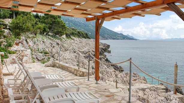 Eine terrasse mit steinbelag, weißen Liegestühlen, einer Holz-Pergola und Blick auf Meer und Berge.