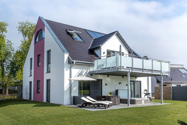 Une maison moderne dotée d’un balcon en verre, d ’une terrasse avec des chaises longues et d’une pelouse bien entretenue.