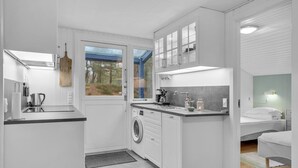 A compact, modern kitchen with a built-in washer and dryer, a large window with a view of trees, and a small bedroom with a bed and nightstand. - 2 bedroom amazing home in Nexø (Nexø)